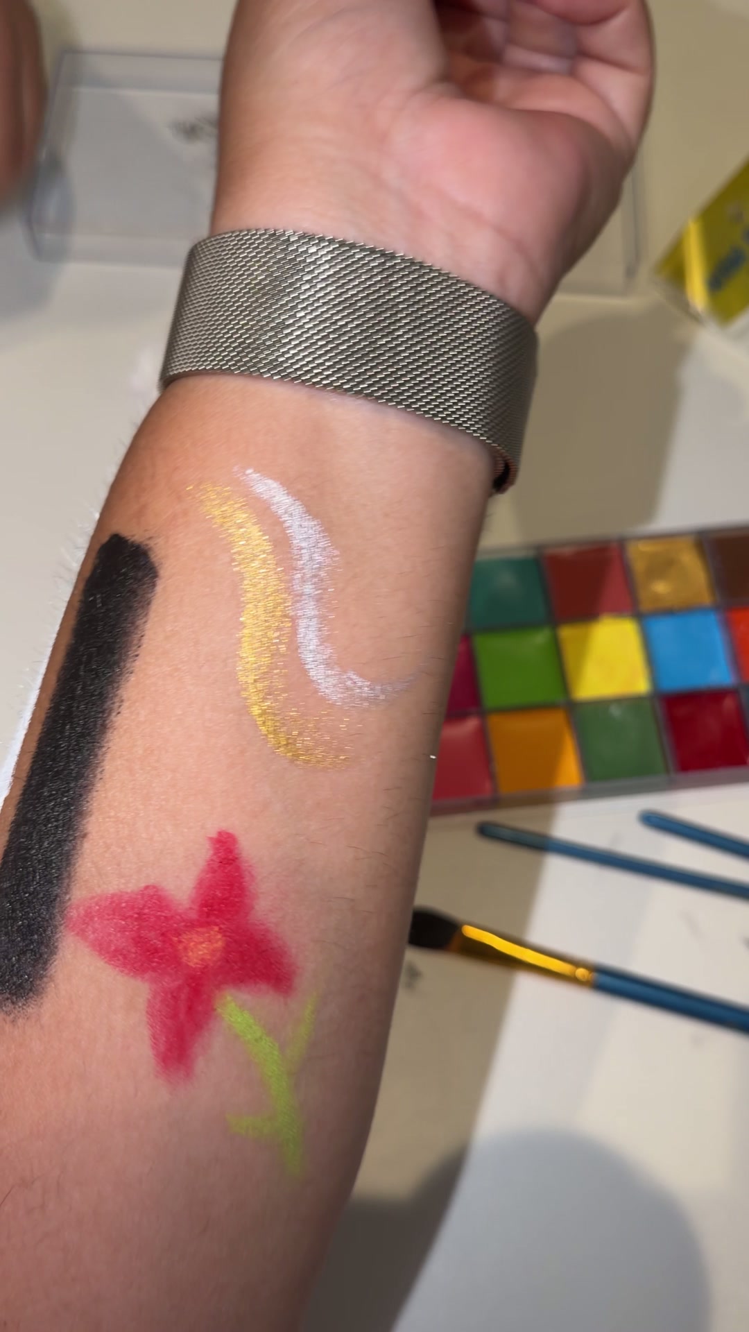 Brazo de Yane con una flor pintada y muestras de negro, dorado y plateado junto a la paleta Athena